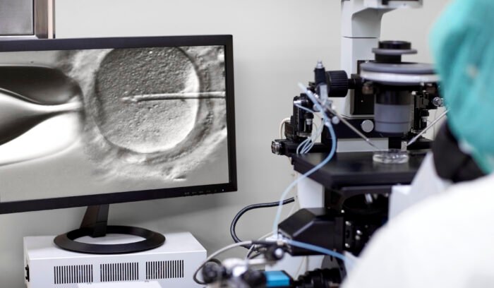 A microscopic image of intracytoplasmic sperm injection (ICSI) displayed on a computer screen, where the needle is in the process of injecting sperm into the egg cell, with laboratory equipment and a microscope visible in the background.